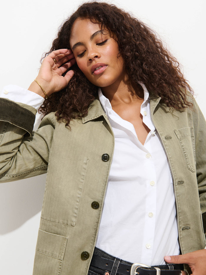 A woman with curly hair wears the Britt Work Jacket in Herringbone, featuring an olive green shade and corduroy collar, over a white shirt. She stands against a white background, eyes closed, tilting her head with one hand near her ear.