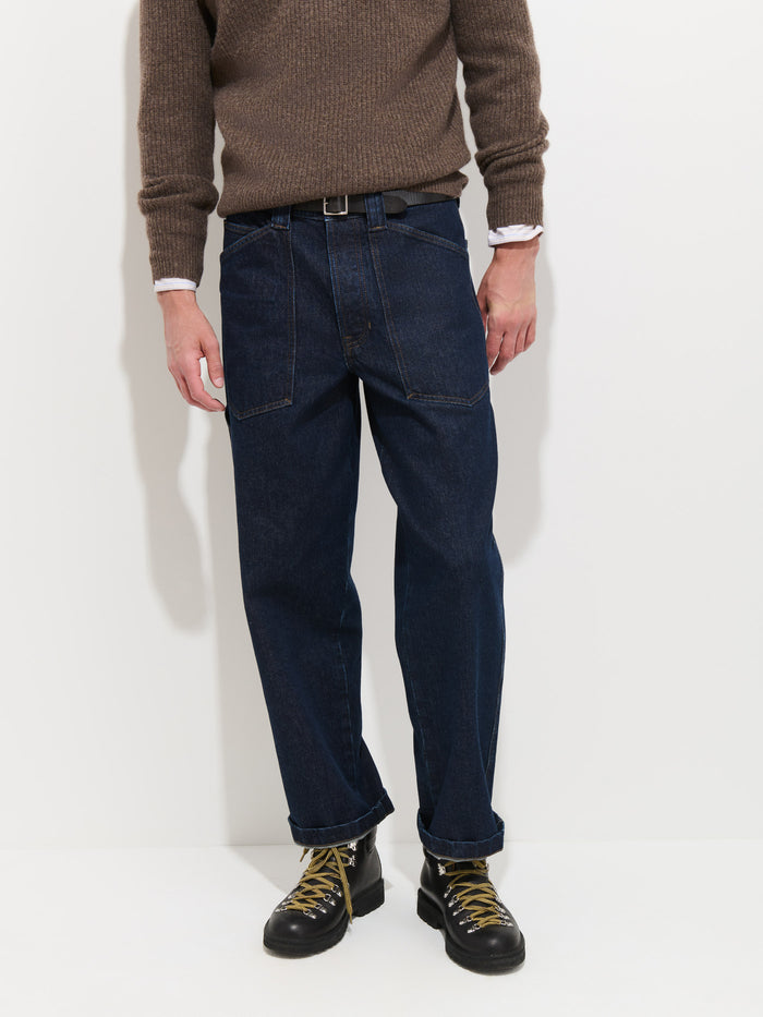 A person stands against a white background, dressed in a brown sweater, white shirt, and the Buckle Back Straight Leg Jean—1940s workwear jeans featuring wide legs and a straight-leg fit. The jeans are paired with black lace-up boots, and the person’s head is not visible in the image.