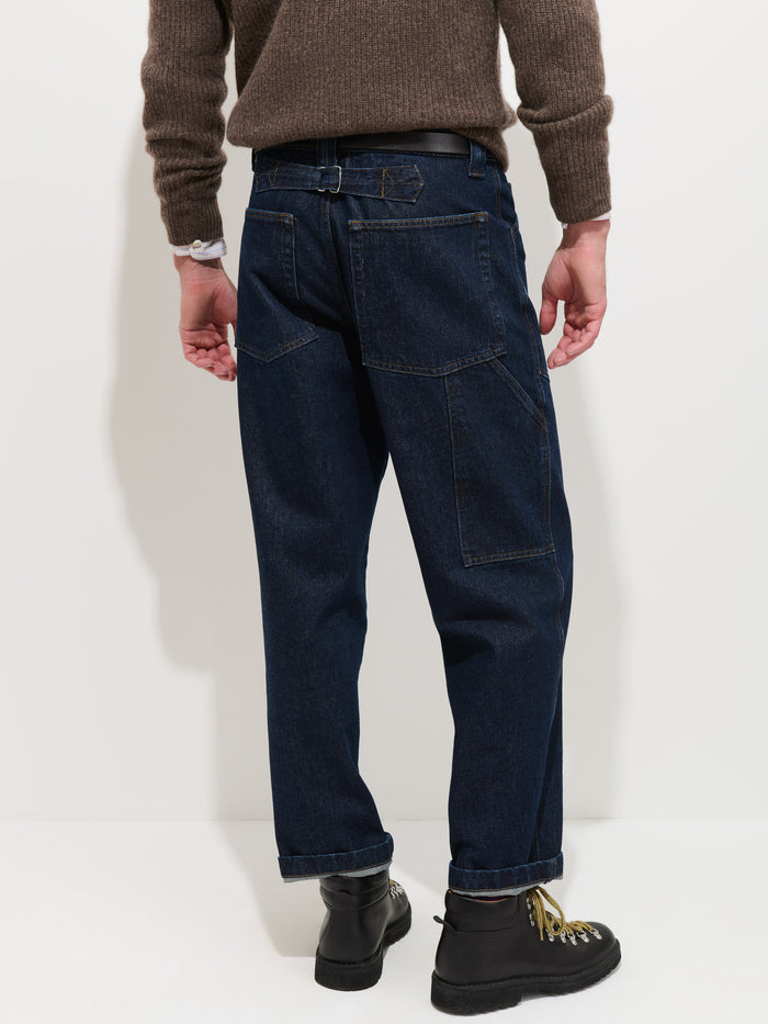 A person wearing the Buckle Back Straight Leg Jean, inspired by 1940s workwear and featuring a dark blue wash with a straight-leg fit, paired with a brown sweater, black boots, and a watch, is shown from the back against a plain white background.