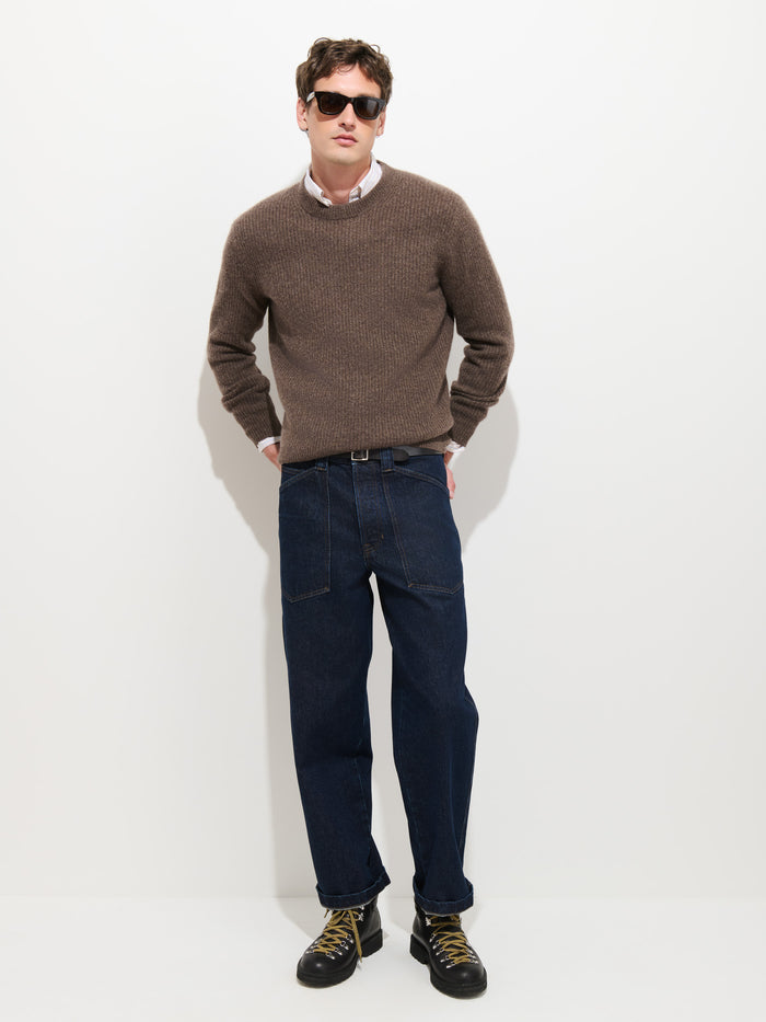A man wearing sunglasses, a brown sweater over a collared shirt, Buckle Back Straight Leg Jeans in a loose, dark blue 1940s-inspired fit, and black shoes with yellow laces stands against a plain white background.