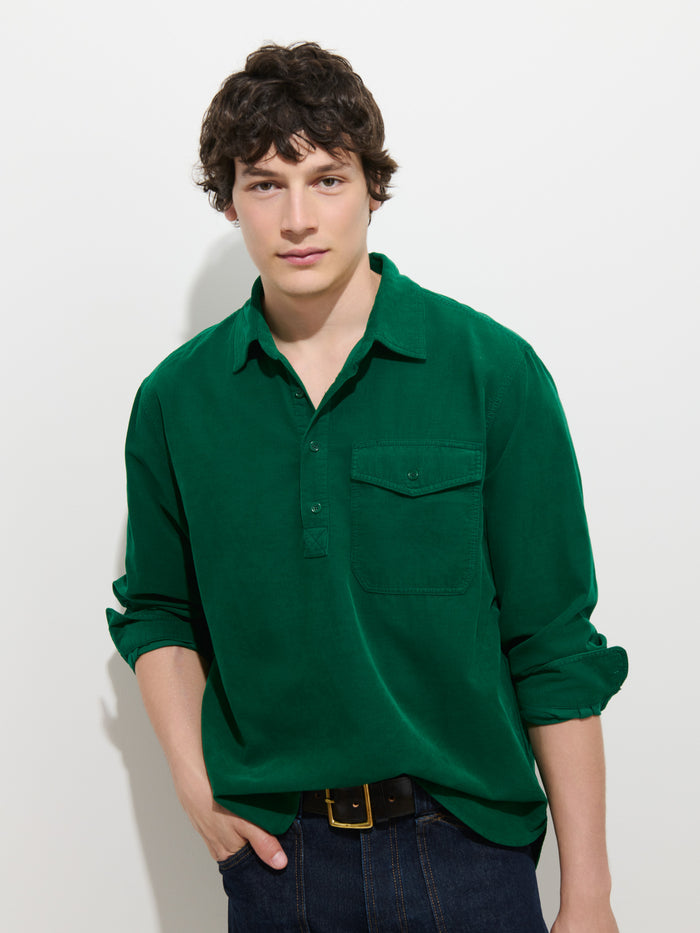A young person with short dark hair wearing the Carter Popover In Fine Wale Corduroy—a green, 100% cotton lightweight corduroy shirt—paired with dark jeans stands against a plain white background, looking at the camera.