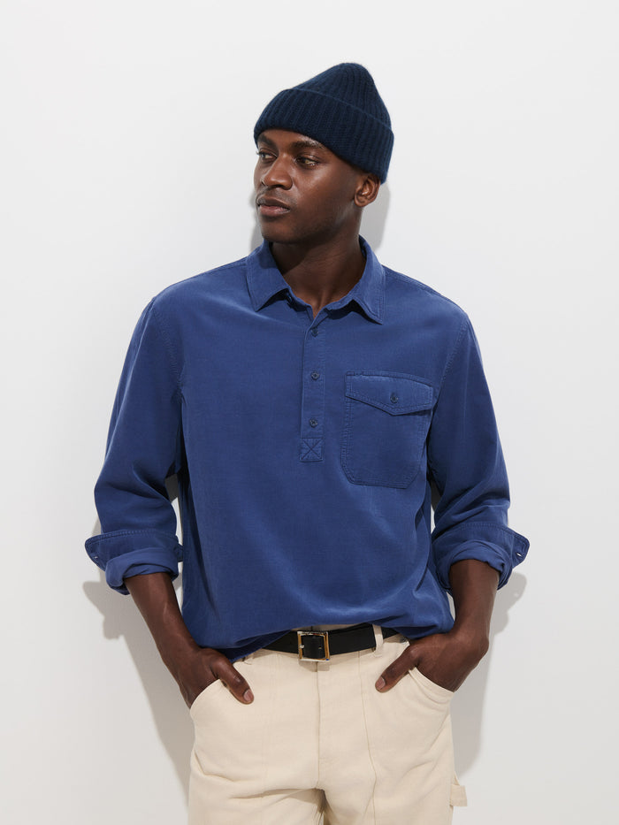A person wearing a navy knit beanie, the Carter Popover In Fine Wale Corduroy—a blue men’s long-sleeve button-up shirt with a chest pocket—and cream pants stands against a plain white background with their hands in their pockets.