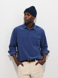 A person wearing a navy knit beanie, the Carter Popover In Fine Wale Corduroy—a blue men’s long-sleeve button-up shirt with a chest pocket—and cream pants stands against a plain white background with their hands in their pockets.