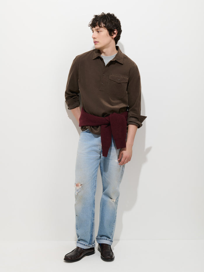 A man stands against a white wall, wearing the Carter Popover in Fine Wale Corduroy—a brown men’s cotton shirt whose lightweight corduroy adds subtle texture to his relaxed look—paired with light ripped jeans, black shoes, and a maroon sweater tied around his waist.