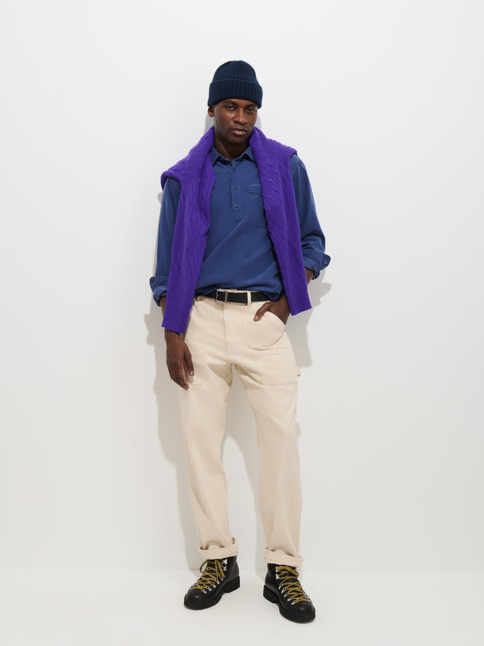 A person wearing a navy beanie, the Carter Popover In Fine Wale Corduroy—an airy blue casual men’s shirt—paired with a purple vest, beige pants, and black boots stands against a white background, looking at the camera with a relaxed pose.