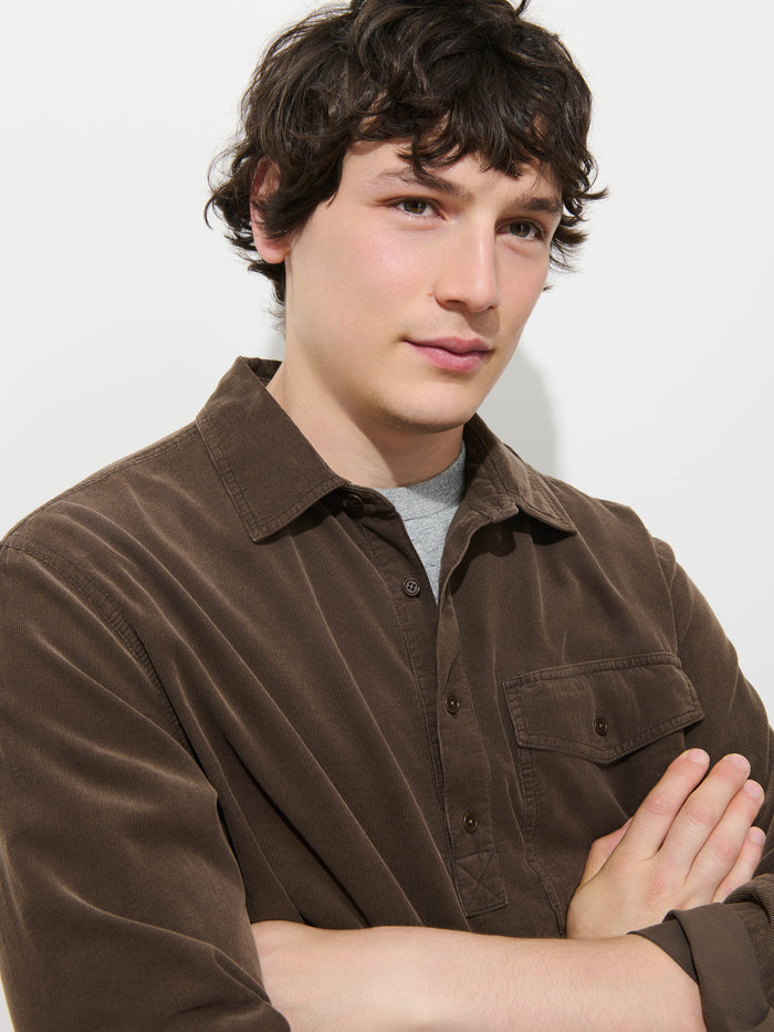 A young man with wavy dark hair, wearing the Carter Popover In Fine Wale Corduroy—a brown men’s shirt styled over a gray t-shirt—stands with his arms crossed and a neutral expression against a plain white background. The lightweight fine wale corduroy fabric adds subtle texture to his relaxed look.