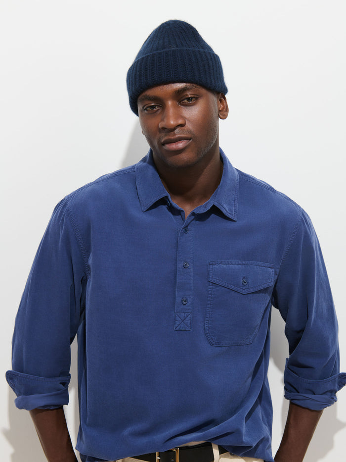 A man wearing a navy beanie and the Carter Popover in Fine Wale Corduroy—a blue men’s casual shirt—stands against a plain white background, looking at the camera with a neutral expression. The lightweight fine wale corduroy fabric of this popover adds subtle texture and comfort to his relaxed style.