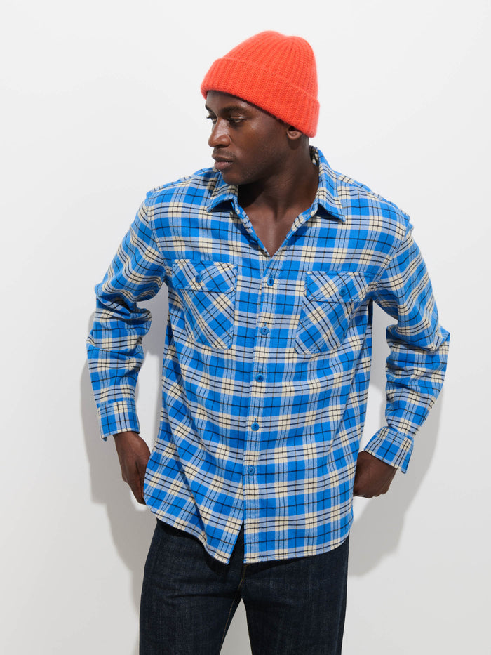 A person stands against a plain white background, looking to the side while wearing the Cashmere Beanie in bright orange, a blue plaid button-up shirt, and dark jeans.
