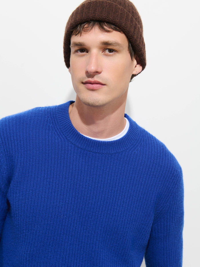 A person models the Cashmere Beanie in soft knit brown, paired with a bright blue sweater, standing against a plain white background and gazing neutrally at the camera.