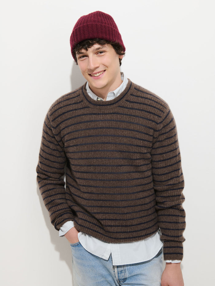 A young man stands against a white background, smiling with one hand in his pocket. He is wearing a brown and black striped sweater, light blue jeans, and the Cashmere Beanie in soft red knit.