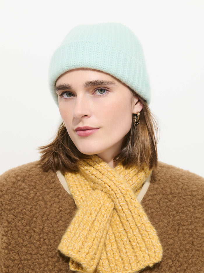 A person wearing the Cashmere Beanie in light blue, gold hoop earrings, a brown sweater, and a yellow soft knit scarf looks at the camera against a plain white background.