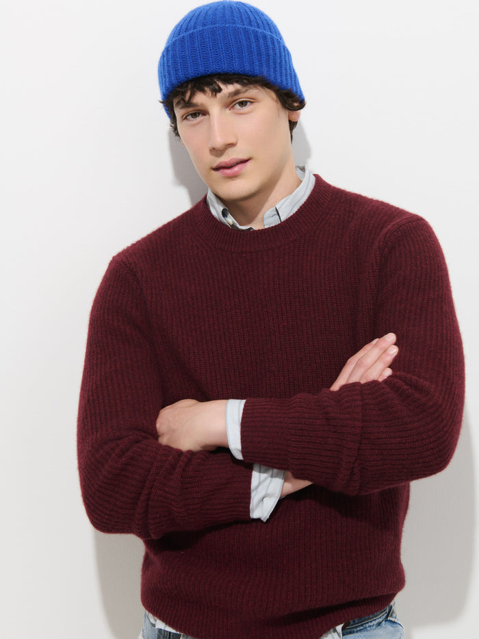 A person wearing a blue Cashmere Beanie, maroon sweater, and light-colored collared shirt stands against a white background with arms crossed, looking at the camera.