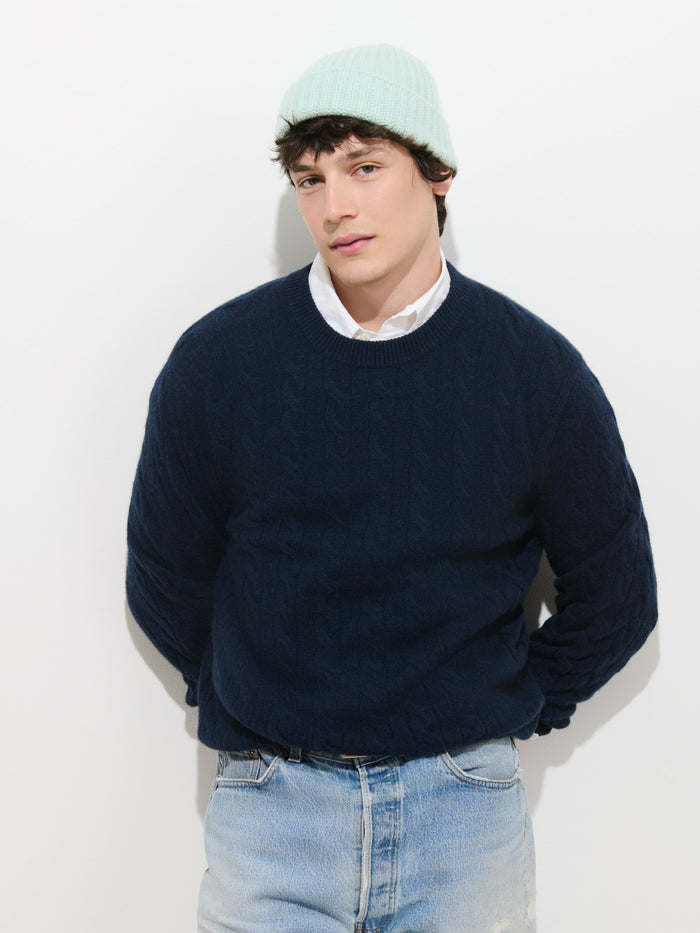 A person wearing the Cashmere Beanie in soft knit light blue, paired with a white collared shirt, a navy blue sweater, and light blue jeans stands against a plain white background, looking at the camera with hands behind their back.