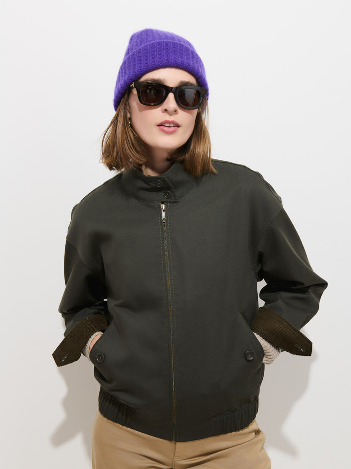 A person wears the Cashmere Beanie in soft knit purple, paired with dark sunglasses, a black zip-up jacket, and beige pants as they stand against a plain white background with hands in their jacket pockets.