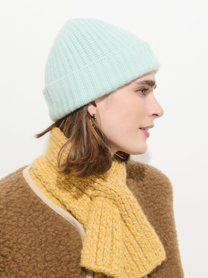A person in profile wears the Cashmere Beanie in light blue, paired with a yellow textured scarf, a brown coat, and small hoop earrings against a plain white background.