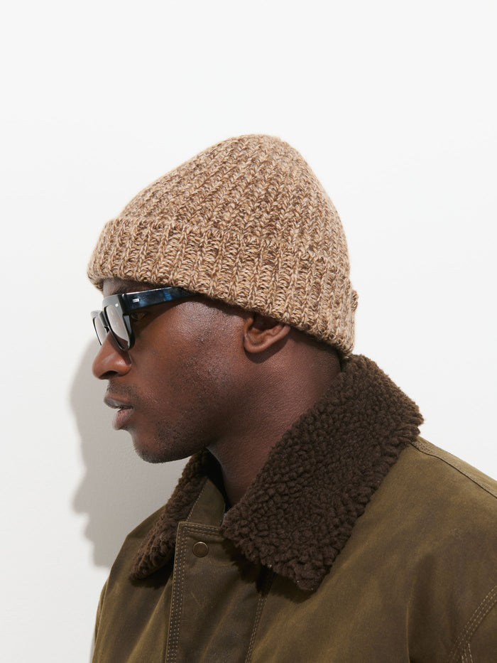 A person in profile view wears the Cashmere Marled Beanie in tan, along with dark sunglasses and a brown jacket with a shearling collar, standing against a plain white background.