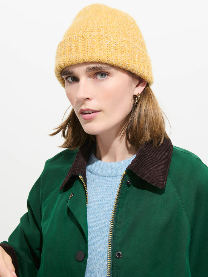 A person with shoulder-length brown hair wears the Cashmere Marled Beanie in yellow, paired with a green jacket featuring a dark brown collar and a light blue sweater, looking slightly to the side against a plain white background.