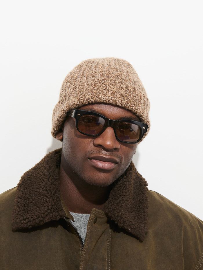 A person wearing dark sunglasses, a Cashmere Marled Beanie in beige, and a brown jacket with a shearling collar stands against a plain white background.