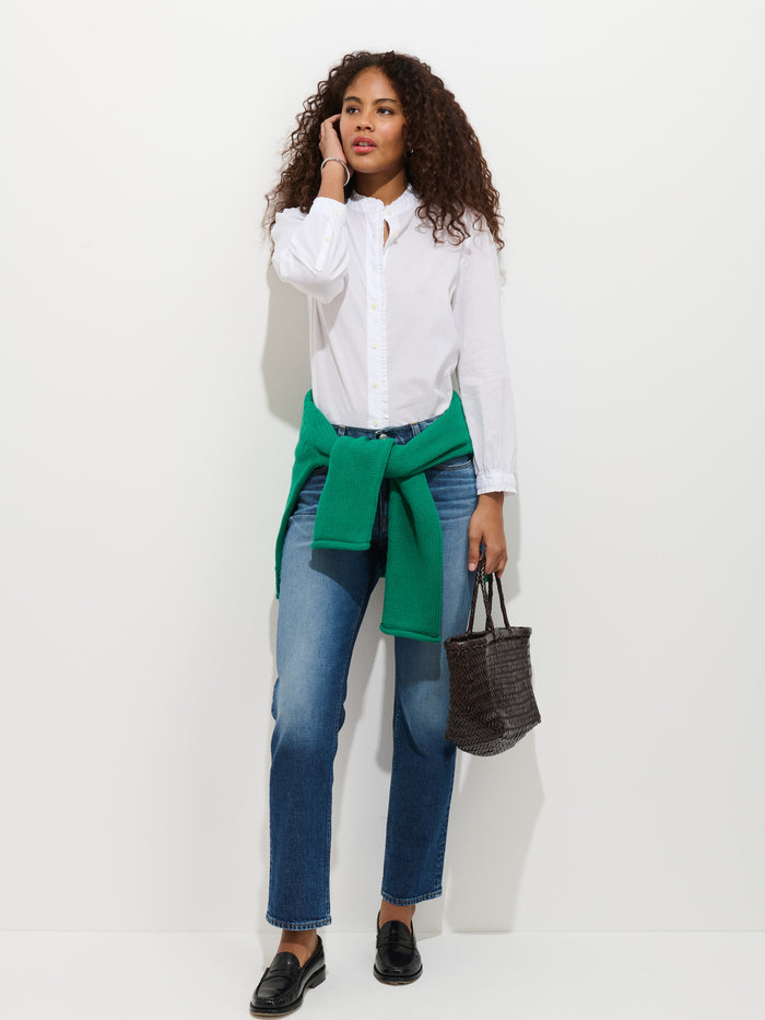 A woman with curly hair channels French style in the Chantilly Ruffle Shirt in Cotton Voile, blue jeans, black loafers, and a green sweater at her waist. She holds a black woven bag and stands before a plain white background.