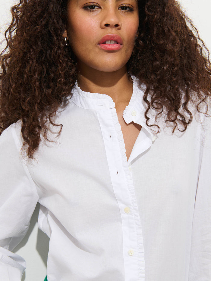 A woman with curly hair stands confidently against a white background, wearing the Chantilly Ruffle Shirt in Cotton Voile—a French-inspired, garment-dyed blouse featuring a ruffled collar.