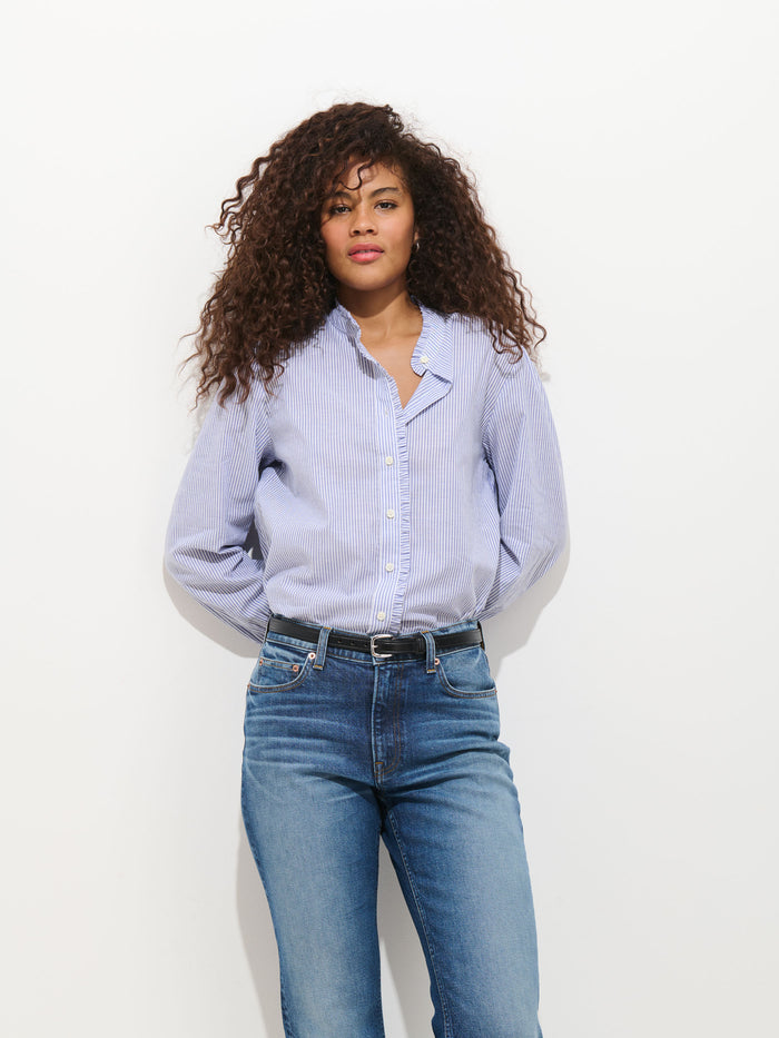 A woman with curly hair stands against a white background wearing the Chantilly Striped Ruffle Shirt In Cotton Voile—this blue and white cotton blouse pairs perfectly with jeans. Her hands are behind her back and she has a neutral expression.