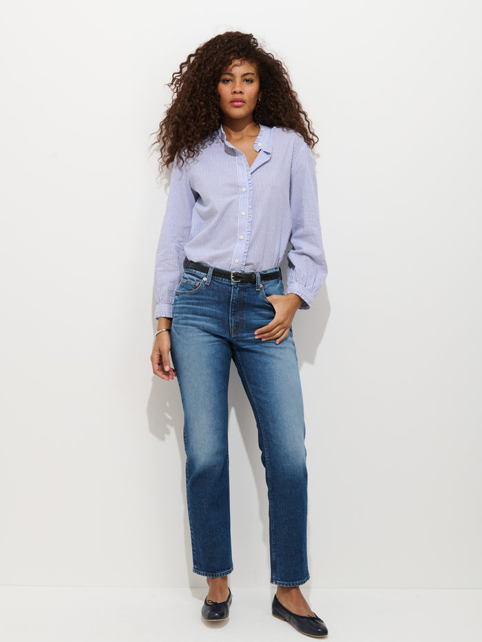 A woman with curly hair stands against a white background, wearing the Chantilly Striped Ruffle Shirt in Cotton Voile with blue jeans, a black belt, and black flats. One hand is in her pocket and she has a relaxed expression.