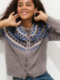 A person with curly hair smiles while wearing the Charlotte Fairisle Sweater in Wool—featuring a gray, blue, white, and black yoke pattern. Their hands rest near their neck; they are casually dressed in jeans.