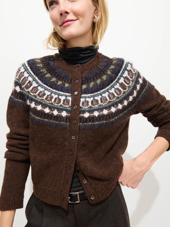 A woman stands against a plain white background, smiling slightly as she wears the Charolette Fairisle Sweater in Wool—a brown cardigan featuring a colorful, vintage-inspired patterned yoke—layered over a black turtleneck.