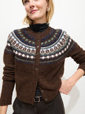 A woman stands against a plain white background, smiling slightly as she wears the Charolette Fairisle Sweater in Wool—a brown cardigan featuring a colorful, vintage-inspired patterned yoke—layered over a black turtleneck.