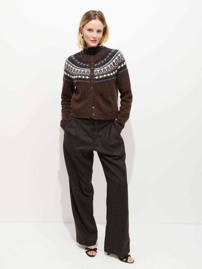 A person stands against a white background wearing the Charolette Fairisle Sweater in Wool—a vintage-inspired, patterned brown cropped knit—paired with wide-leg dark pants and black sandals, hands tucked into their pockets and a relaxed expression on their face.