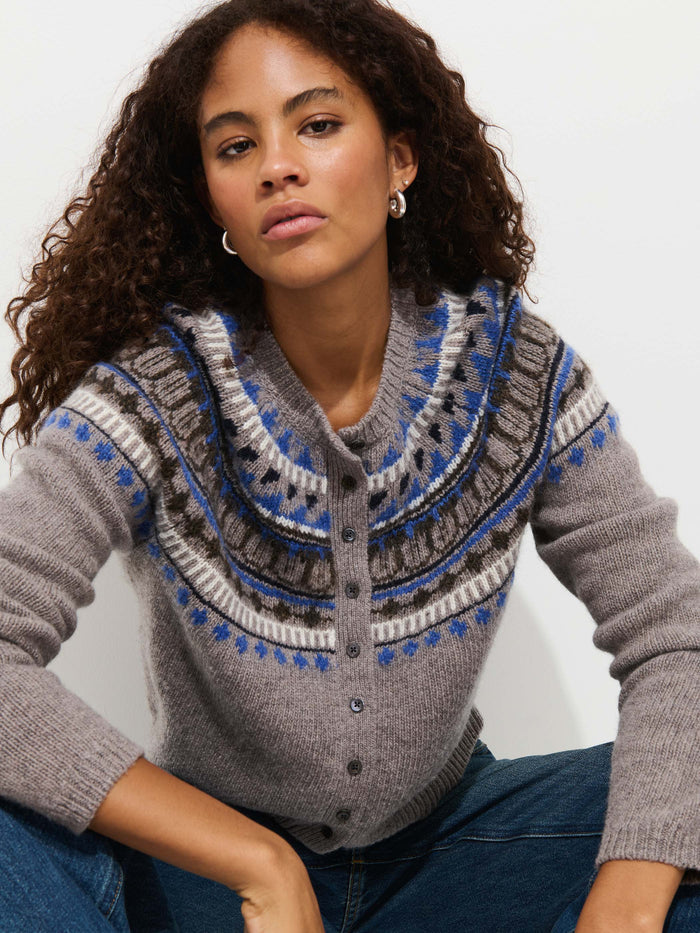 A woman with curly hair models the Charlotte Fairisle Sweater in Wool, featuring a blue, white, and brown vintage Fair Isle yoke. She sits against a white background, looking directly at the camera with a neutral expression.
