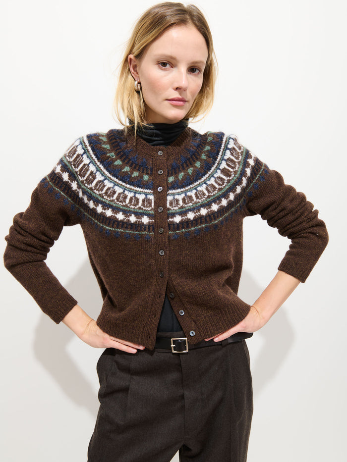 A woman stands with her hands on her hips against a plain white background, styled in the Charolette Fairisle Sweater in Wool—a vintage-inspired piece featuring a brown merino mohair blend with multicolored patterns—layered over a black turtleneck and paired with dark trousers.