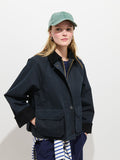A woman models the Chiltern Cropped Jacket In Waxed Cotton—featuring a corduroy collar—with a green baseball cap, hoop earrings, and a striped garment tied at her waist, all set against a plain white background.
