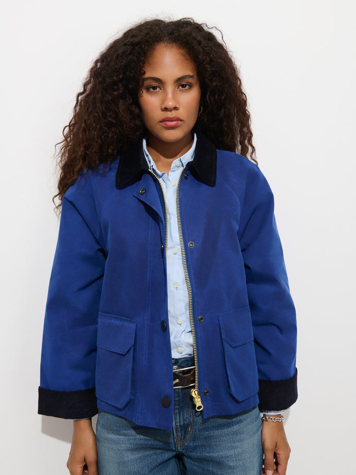 Wearing the Chiltern Cropped Jacket in Waxed Canvas in blue over a light blue shirt and jeans, a person with long curly hair stands against a plain white background with a neutral expression.