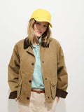 A person in a yellow baseball cap, green and white striped shirt, beige pants, and the Chiltern Cropped Jacket In Waxed Canvas with a dark collar stands against a plain white background.