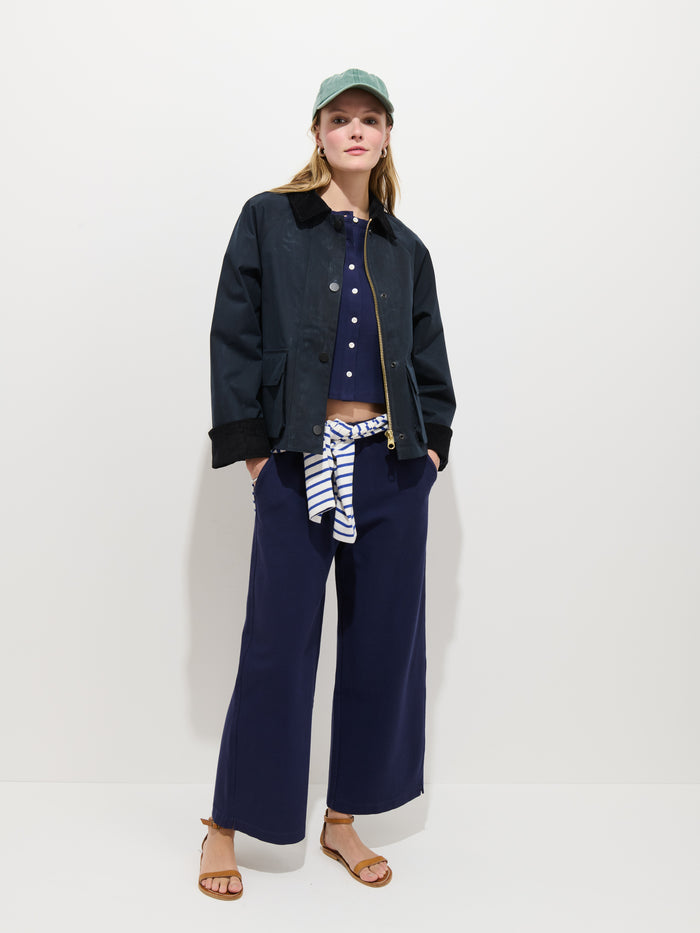 A woman stands against a white background wearing the Chiltern Cropped Jacket In Waxed Cotton with a dark corduroy collar, green cap, navy top, wide-leg pants, sandals, and a striped shirt tied at her waist. She looks confidently at the camera.