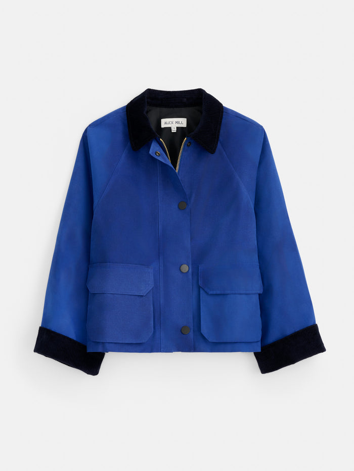 Introducing the Chiltern Cropped Jacket In Waxed Canvas—a blue cropped jacket featuring a black collar and cuffs, front button closure, and two spacious front pockets. Designed with a waxed cotton canvas for enhanced durability and modern style, this women’s outerwear piece displays an interior ALEX MILL label for a signature touch.