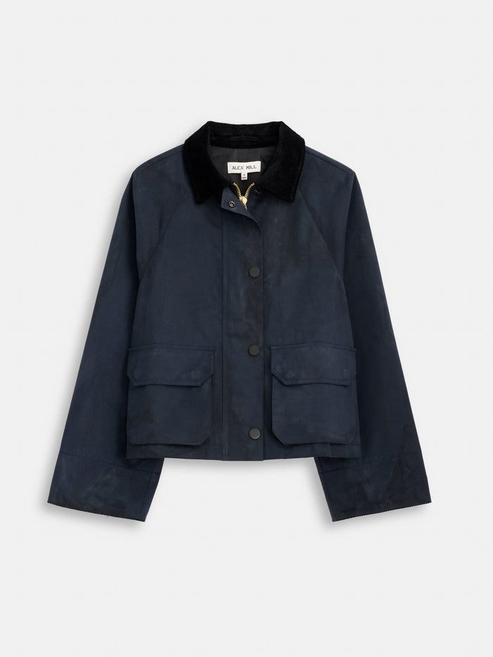 The Chiltern Cropped Jacket In Waxed Cotton is a women’s dark navy blue outerwear with a black corduroy collar, front snap buttons, two large flap pockets, long sleeves, and a cropped boxy fit, shown on a plain white background.