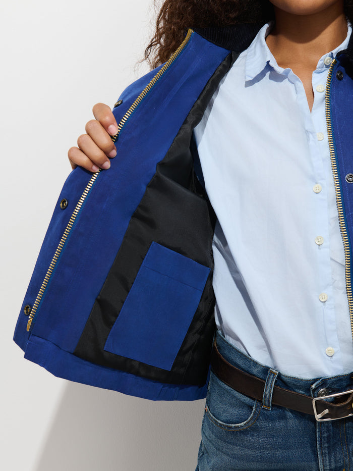 A person in a light blue shirt and jeans opens the Chiltern Cropped Jacket in Waxed Canvas, revealing its black inner lining and a small interior pocket. The person’s face is partially out of frame.