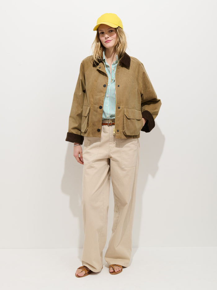 A woman stands against a white background, wearing a yellow cap, blue striped shirt, beige wide-leg pants, brown sandals, and the Chiltern Cropped Jacket in Waxed Canvas with dark cuffs and collar.
