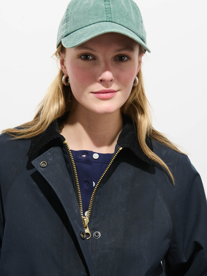 A person with long blond hair wearing a green baseball cap, gold hoop earrings, a navy top, and the Chiltern Cropped Jacket In Waxed Cotton stands against a plain white background, looking directly at the camera.