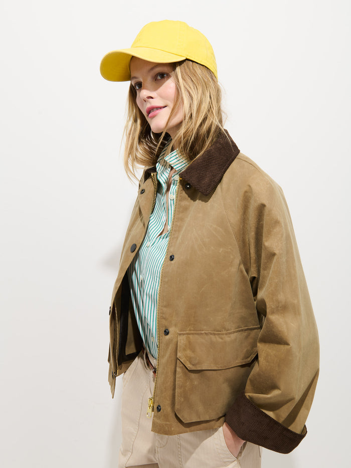 A woman in a yellow cap, blue and white striped shirt, light pants, and the Chiltern Cropped Jacket In Waxed Canvas stands against a plain white background—a modern update on women’s outerwear.