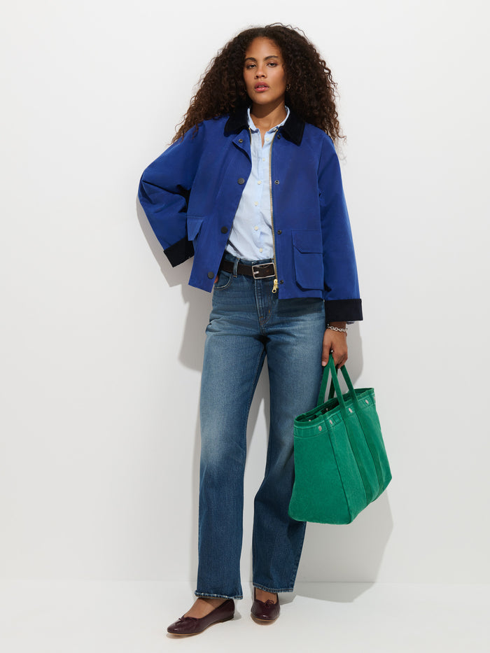 A woman with curly hair stands against a white background wearing the Chiltern Cropped Jacket in Waxed Canvas, paired with a light blue shirt, jeans, brown flats, and holding a large green tote bag.