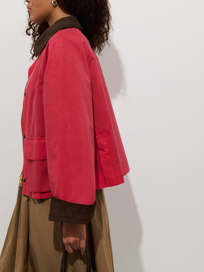 Wearing the Chiltern Cropped Jacket in Waxed Canvas over a brown shirt and tan skirt, a person with curly hair stands against a white background, holding a woven bag. The image is cropped at the shoulders and knees.