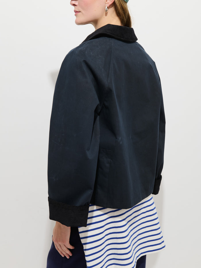 A person wearing the Chiltern Cropped Jacket in waxed cotton, featuring a black corduroy collar and cuffs over a blue-and-white striped shirt, stands against a plain white background facing away and slightly to the side.