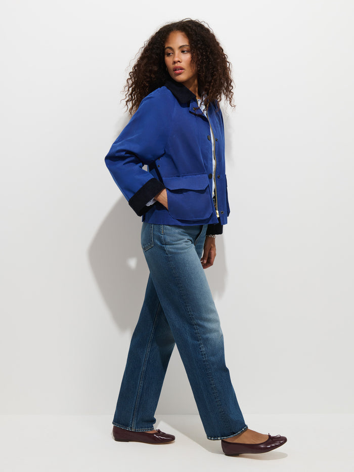 A person with curly hair, wearing the Chiltern Cropped Jacket in Waxed Canvas—a sophisticated piece of women’s outerwear in blue—paired with jeans and brown flats, stands in profile against a plain white background, gazing forward with one hand tucked into a pocket.