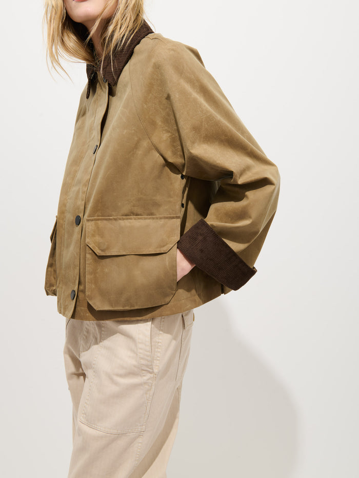 A person models the Chiltern Cropped Jacket in Waxed Canvas—a light brown, boxy women’s jacket with dark brown collar and cuffs—paired with beige pants, standing against a plain white background.