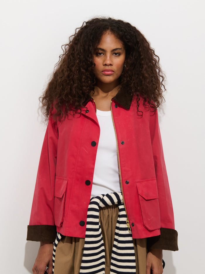 A woman with long, curly hair wears the Chiltern Cropped Jacket in waxed canvas over a white top and tan pants, with a striped sweater tied at her waist. She stands against a plain white background, looking directly at the camera.