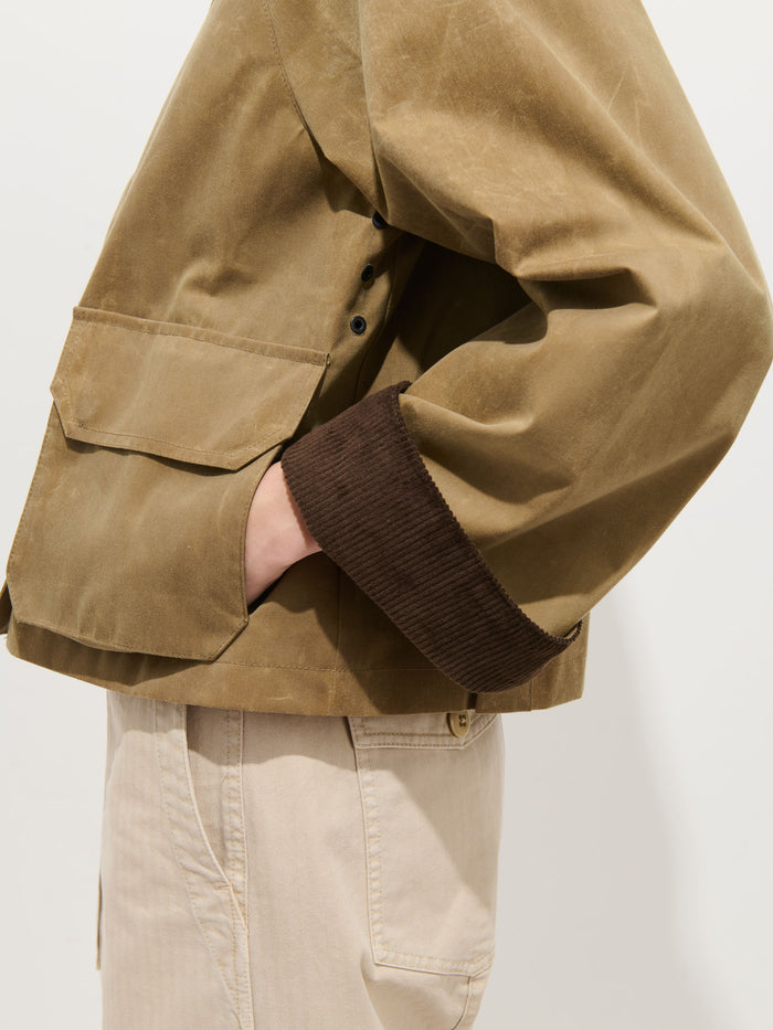 A person wears the Chiltern Cropped Jacket in waxed canvas with brown corduroy cuffs, their left hand in the pocket. Light beige pants complete the look. The torso and arm are highlighted to showcase stylish women's outerwear details.
