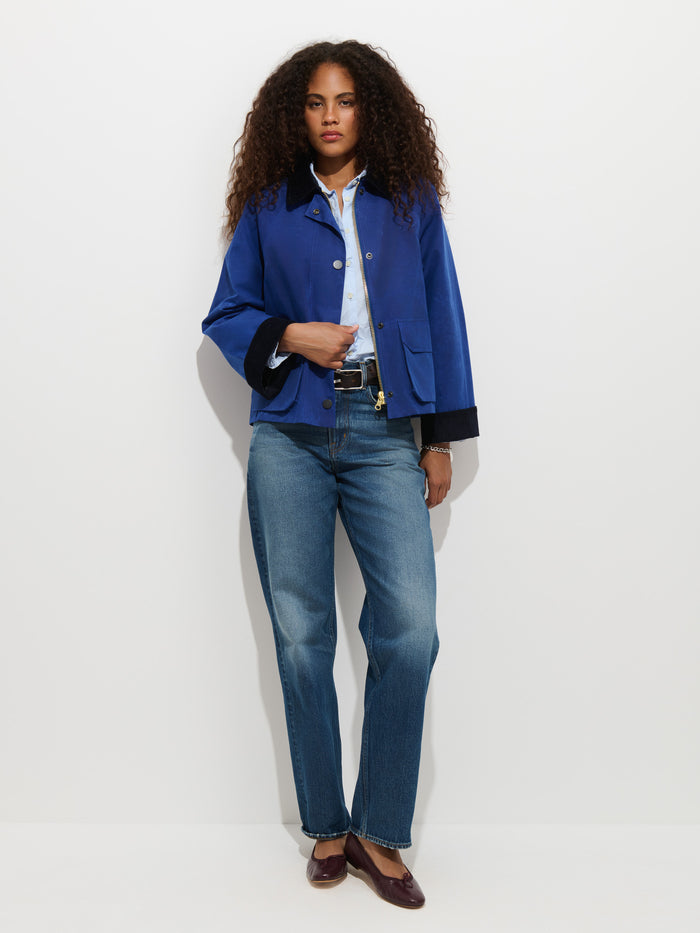 A woman with curly hair stands against a white background, wearing the Chiltern Cropped Jacket in Waxed Canvas over a light shirt, paired with blue jeans and maroon flats.